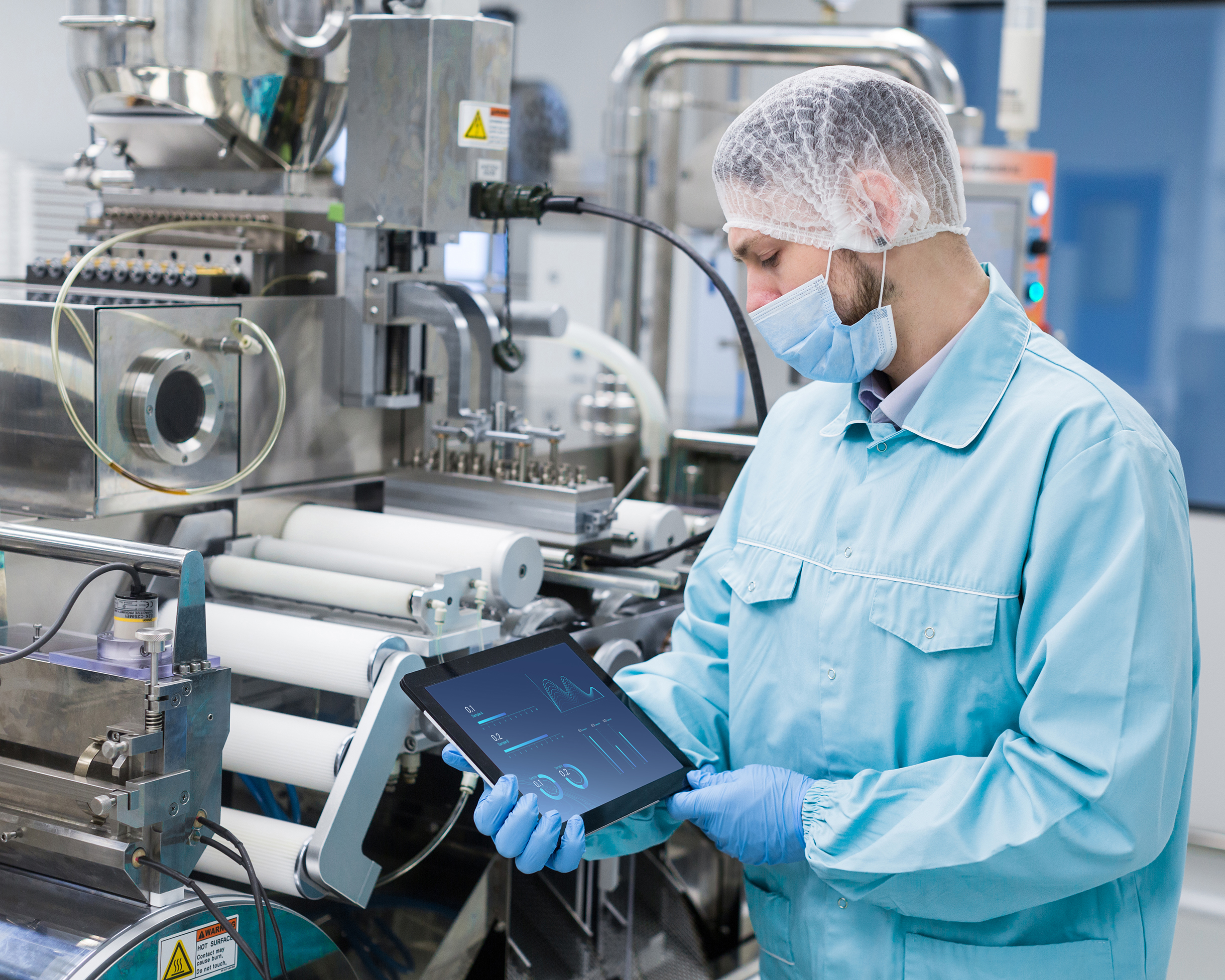 caucasian scientist man in blue lab uniform stand near manufacture machine with shafts, show empty tablet, close picture, focus on tablet, look at tablet